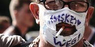 Mann mit Mund/Nasenmaske, wobei der Mund durch ein aufgerissenes Loch in der Maske schaut - in ihm steckt eine Zigarette
