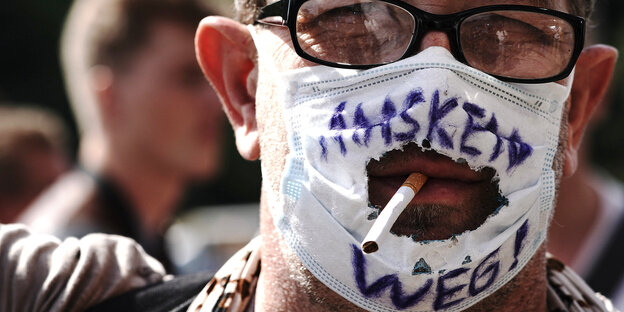 Mann mit Mund/Nasenmaske, wobei der Mund durch ein aufgerissenes Loch in der Maske schaut - in ihm steckt eine Zigarette