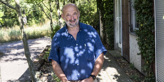 Fred Schöner hat kurzes graues Haar und einen kurz geschnittenen grauen Bart. Er trägt ein leuchtend blaues Leinenhemd mit kurzen Ärmeln und steht im schattigen Garten der Hellersdorfer Kultureinrichtung "Kiste".