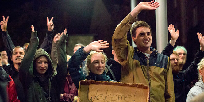 Menschen winken, eine Frau hält ein Pappschild mit der Aufschrift "Welcome"