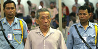Kaing Khek Iev steht zwischen zwei Polizisten