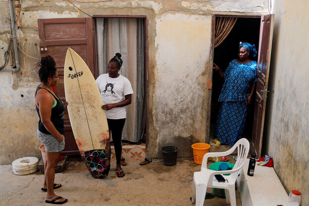 Eine Frau schaut aus der Tür auf zwei Frauen neben einem weißen Surfbrett