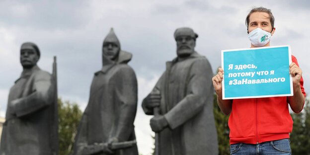 Ein junger Mann steht mit eiem Schild und Gesichtsmaske in Nowosibirsk vor drei sowjetischen Heldenstatuen