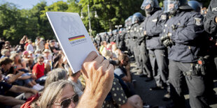 Menschen sitzen auf der Straße einer Reihe Polizist*innen in Kampfmontur gegenüber. Einer hält das Grundgesetz hoch.