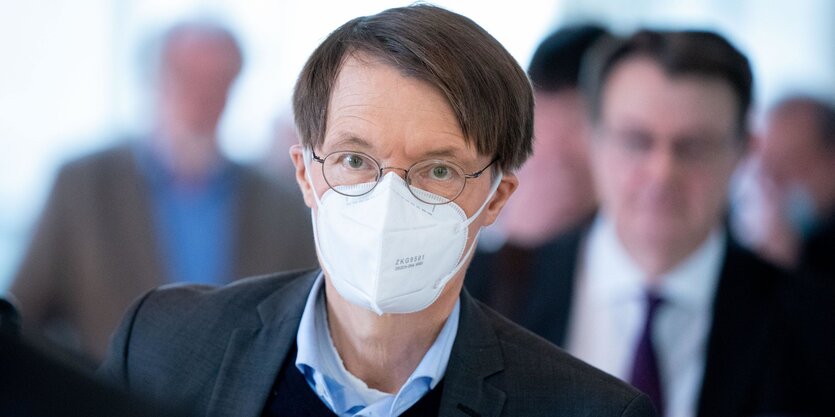 Karl Lauterbach trägt eine Gesichtsmaske, im Hintergrund stehen unscharf einige Menschen.