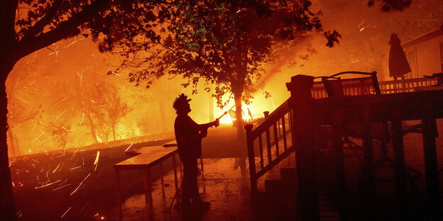 Ein Mann steht mit einem Gartenschlauch vor seinem Haus, im Hintergrund brennt es