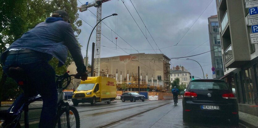 Ein Radfahrer fährt auf einer Straße mit Gleisen an einem parkenden Auot vorbei, im Hintergrund eine Baustelle mit einem Kran