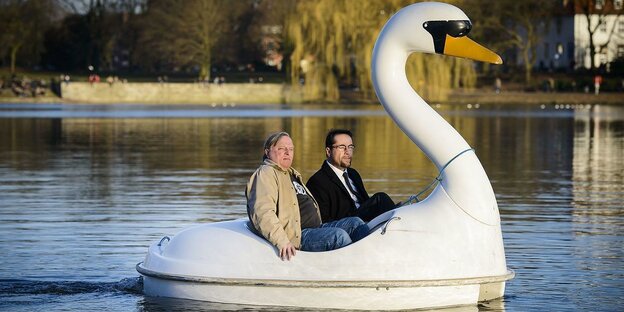 Die Schauspieler Axel Prahl und Jan Josef Liefers sitzen in einem Tretboot, das die Form eines Schwans hat. Das Tretboot ist auf dem Wasser. Prahl ist klein und dick, Liefers ist größer. Beide sind ältere Männer.
