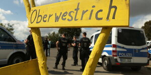 Der Ortsname "Oberwestrich" handgeschrieben auf gelb angemaltem Holz, dahinter Polizisten mit Fahrzeugen