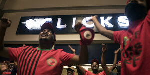 Menschenin roten T-shirts heben die Hände, im Hintergrund ist der Neon-Schriftzug der südafrikanischen Drogeriekette Clicks zu sehen