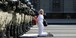Ein Frau, weiß gekleidet und mit einem roten Tuch in den Händen, kniet vor einer Reihe behelmter und mit Tränengasgwehre ausgerüsteter Polizisten