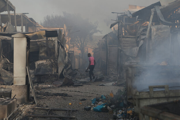 Ein Mensch in einem rosafarbenem T-shirt zwischen ausgebrannten Hütten aus denen Rauch aufsteigt