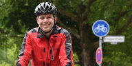 Der Antwalt Andreas Volkmann lächelnd auf seinem Fahrrad, mit Regenjacke und Fahradhelm. Im Hintergrund ein Fahrradweg-Schild.