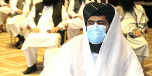 Ein Mann im weißen Gewand und mit Turban trägt einen Mund/Nasenschutz, dahinter sitzen mehrere ähnlich gekleidete Männer