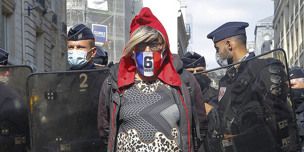 Eine Frau mit roter Kapuze und Tricolore-Mundschutz steht mit dem Rücken vor einem Spalier von Polizisten