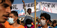 Demonstranten mit Schutzmasken, in der Bildmitte ein Kind auf den Schultern eines Mannes, es streckt den rechten Arm in die Höhe, dahinter ein weißes Transparent mit Aufschrift