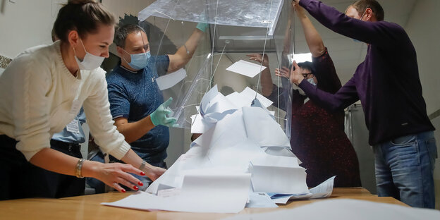 Vier Menschen schütten eine große durchsichtige Wahlurne aus, so dass Stimmzettel auf einen Tisch flattern.