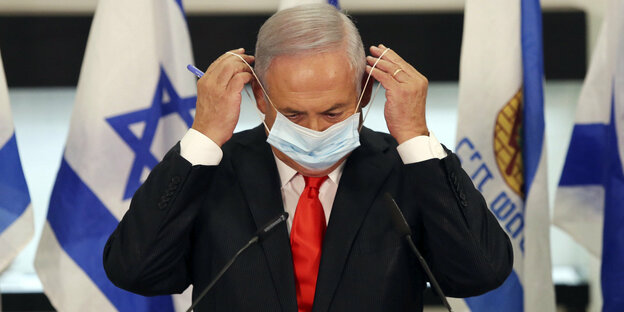 Israels Ministerpräsident Netanjahu setzt sich einen Mundschutz auf, im Hintergrund israelische Flaggen