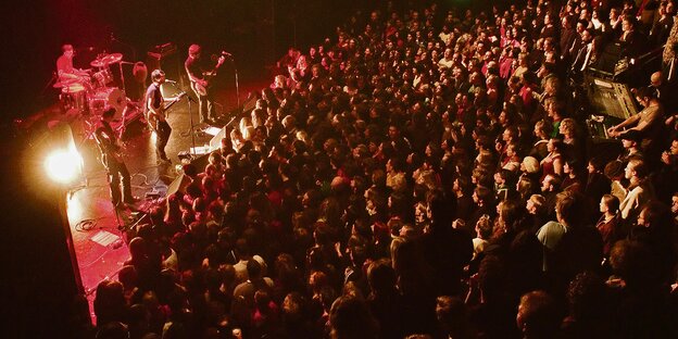 Eine Band spielt in einer voll besetzten Halle mit aufsteigenden Zuschauerrängen.