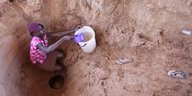 Ruanderin sitzt in einem grob ausgehobenem Brunnen und schöpft mit einem Plastikeimer Wasser.
