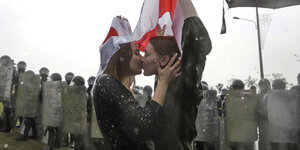 Zwei Demonstrantinnen küssen sich vor einer Riege von Bereitschaftspolizisten, bedeckt von einer ehemaligen belarussischen Nationalflagge während einer Protestkundgebung