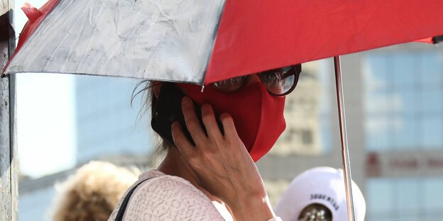 eine Frau mit Schirm und roter Maske telefoniert während einer Demo mit dem Smartphone