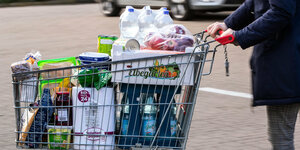 Ein Einkaufswagen mit Lebensmitteln und Produkten des täglichen Bedarfs wird über den Parkplatz eines Supermarkts geschoben