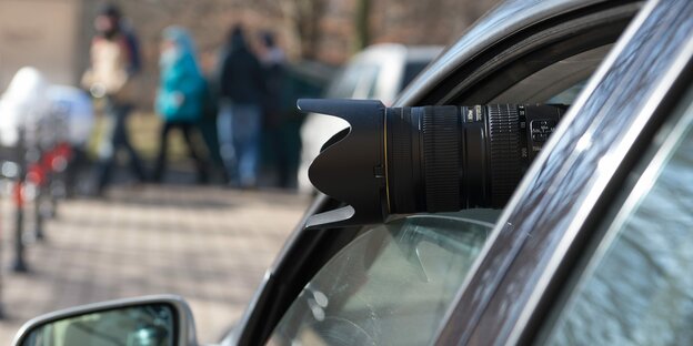 Das Teleobjektiv einer Fotokamera ragt aus dem Seitenfenster eines geparkten Autos.
