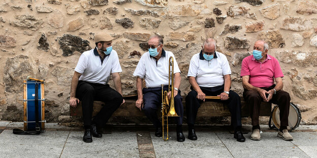 Mitglieder einer lokalen Musikgruppe tragen Mundschutze und sitzen mit ihren Instrumenten auf einer Bank