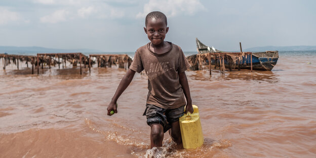 Ein Junge trägt einen gelben Eimer Wasser und läuft bis zu den Knien in bräunlichem, verdreckten Wasser eines Sees