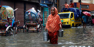 Eine Frau im Sari watet mit zwei Eimern in der Hand auf einer überfluteten Straße