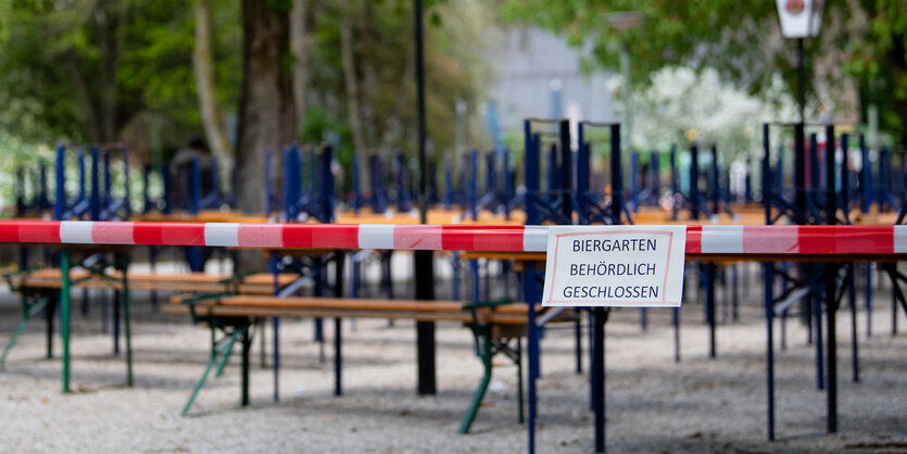 Absperrflatterband mit Corona-Warnschild vor verregneten Bierbänken