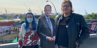 Eine Frau, eine Pappfigur von Friedrich Merz und ein Mann am Hafen