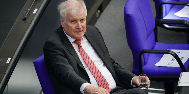 Horst Seehofer sitzt auf einem blauen Stuhl im Bundestag und hält ein Wasserglas.