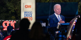 Autos parken vor einem Monitor, auf dem eine Rede von Joe Biden übertragen wird.