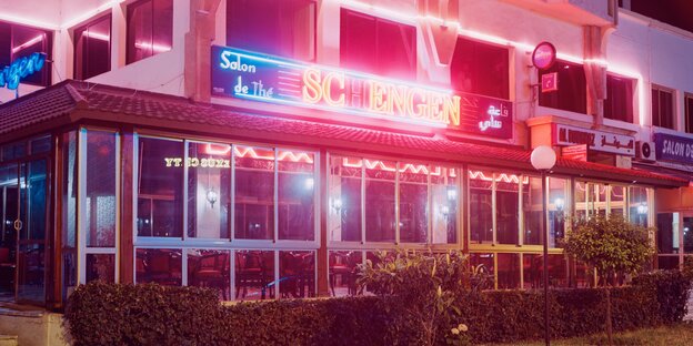Man sieht ein Café in der Nacht, darüber steht "Schengen" in lateinischen Buchstaben, weitere Schriftzeichen sind arabisch