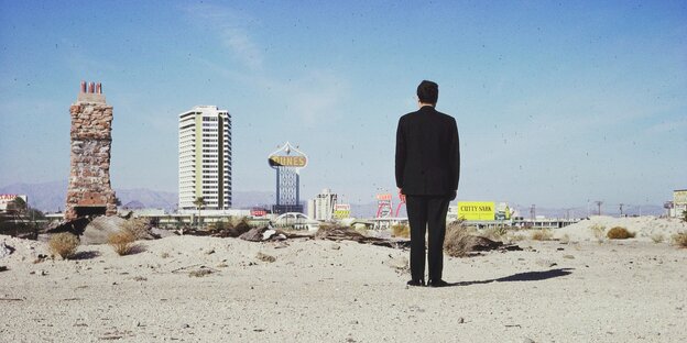 Rückenansicht eines Mannes im Anzug in der Wüste, er blickt auf die Hochhäuser einer Stadt