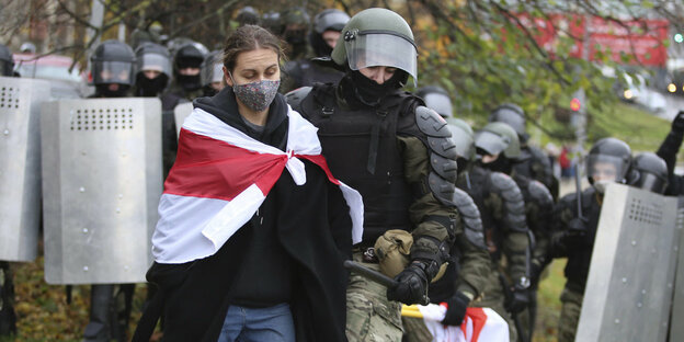 Eine Frau mit einer umeghängten weiß rot gestreiften Flagge wird von einem Polizist in Kampfmontur festgenommen