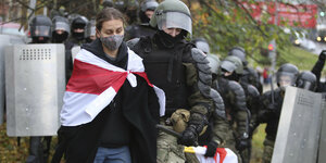 Frau mit Maske und umgehängter weiß-rot-weißer Fahne wird von schwarzgekleideten Sicherheitskräften abgeführt