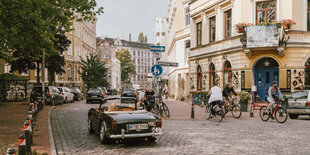 Ein altes Cabrio fährt durch eine Straße mit Koopfsteinfplaster, links Bäume, rechts Altbauteen