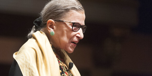 Ruth Bader Ginsburg, seitlich fotografiert, sie trägt einen gelben Schal um die Schultern.