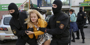 Zwei schwarz maskierte und gekleidete Polizisten nehmen eine Frau mit blonden Haaren und Glitzerrock brutal in die Zange