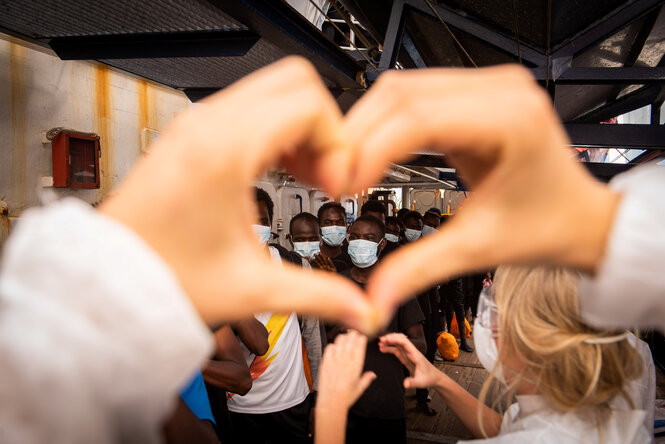 Ein Mitglied der Sea-Watch-Crew formt mit den Händen ein Herz, im Hintergrund verlassen afrikanische Migrant:innen das Schiff Sea Watch 4