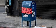 Ein bleuer Briefkasten auf einer Strasse, im Hintergrund ein öffentlicher Fernsprecher