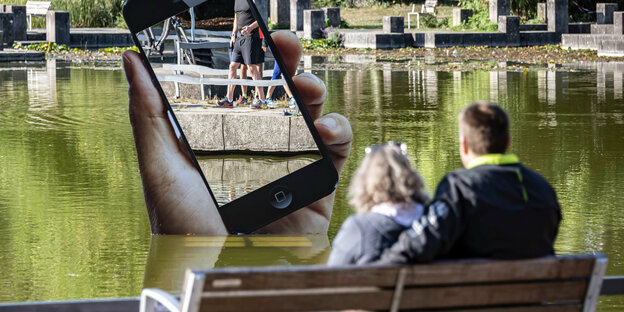 Ein Paar sitzt an einem See, aus dem ein Handy herausragt.