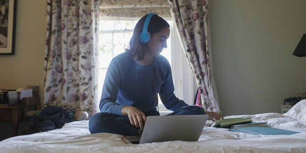 Ein Mädchen sitzt auf einem Bett, sie trägt Kopfhörer, vor ihr ein Laptop