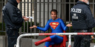 Ein Mann im Supermann-Kostüm sitzt ohne Mundnasenschutz auf einer Bank und wird von 2 Polizisten kontrolliert
