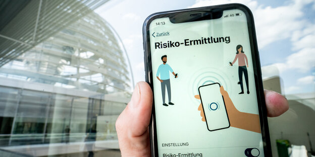 Die Corona-Warn-App mit der Seite zur Risiko-Ermittlung ist im Display eines Smartphone vor der Kuppel des Reichstagsgebäudes zu sehen.
