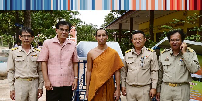 fünf Männer vor einem thailändischen Dorf, der mittlere trägt ein goldenes Mönchsgewand, die anderen sind uniformiert