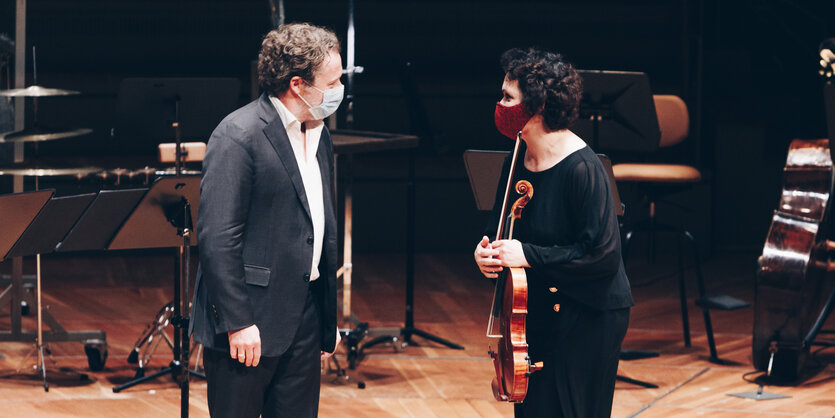 Eine Frau mit Instrument und ein Herr mit Anzug, beide tragen eine Maske, wenden sich auf einer Bühne einander zu.